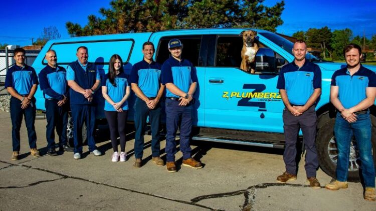 The Z Plumberz of Downriver plumbing team posing in front of their bright blue company truck, with a friendly brown dog sitting in the truck bed.