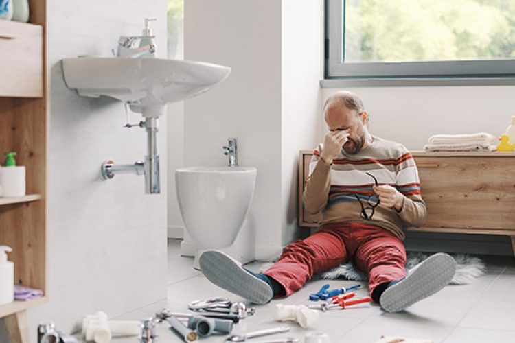 Frustrated homeowner sitting on bathroom floor surrounded by plumbing tools and pipe after failing to fix sink.
