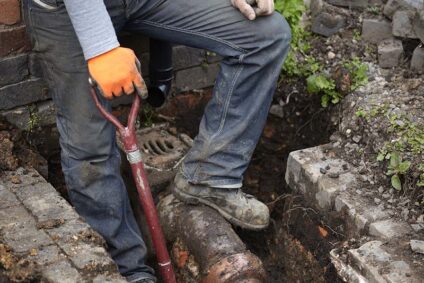 Plumber holding shovel and standing on a partially dug out sewer line.